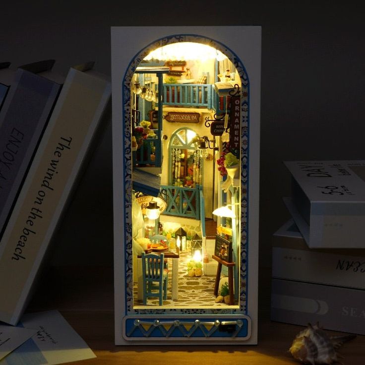 a miniature dollhouse is lit up in the dark with its lights on and books beside it