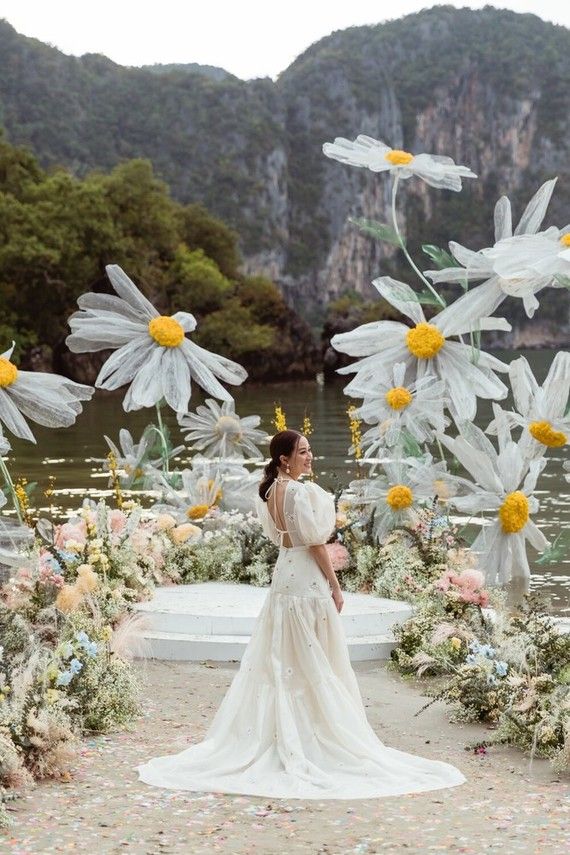 a woman is standing in front of flowers on the ground with mountains in the background