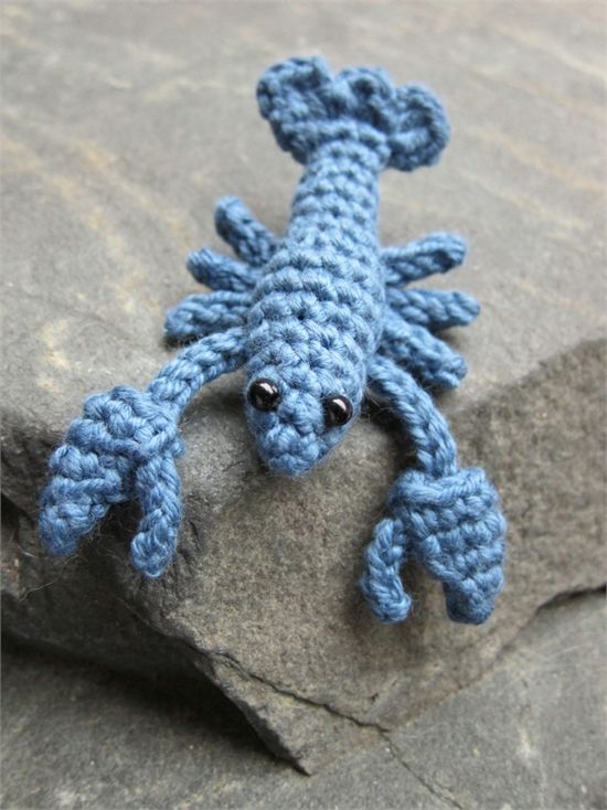 a small crocheted blue gecko sitting on top of a rock next to some rocks