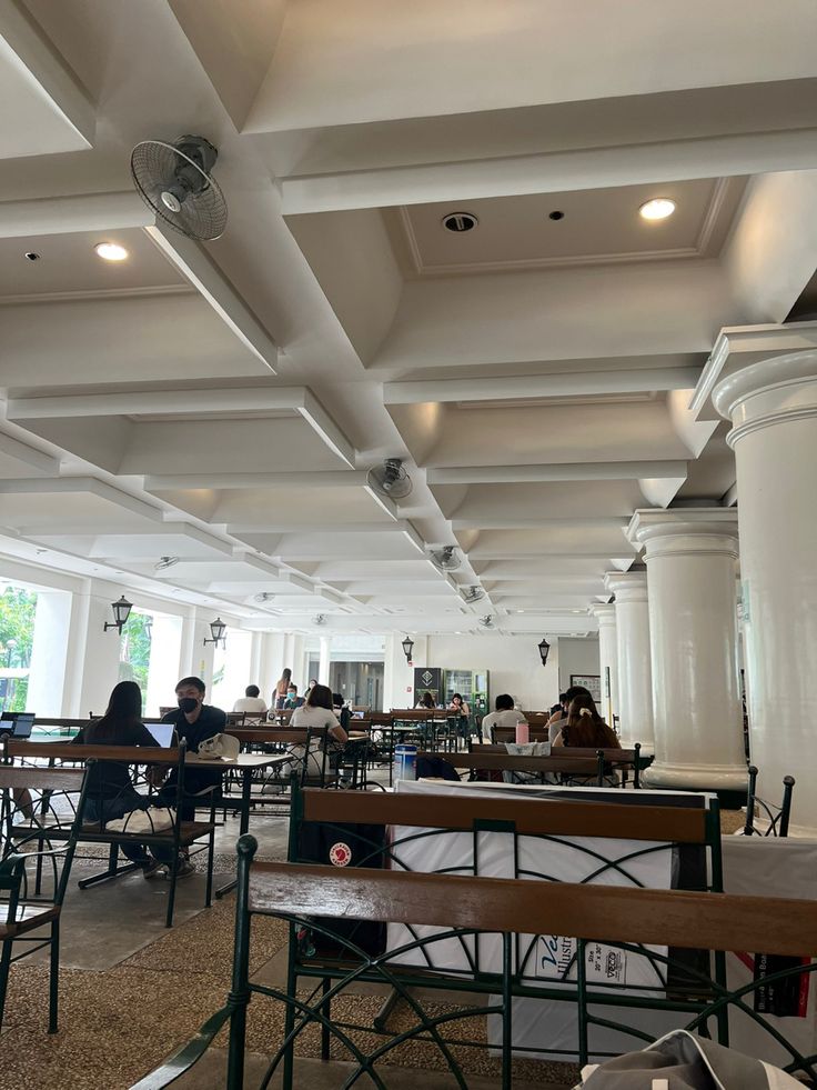 people are sitting at tables in a large room with white walls and ceiling beams, while others sit on their laptops