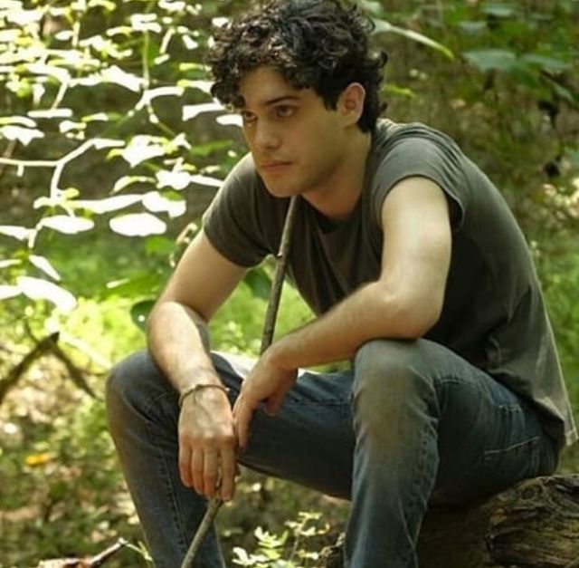 a young man sitting on top of a tree stump holding a stick in his hand