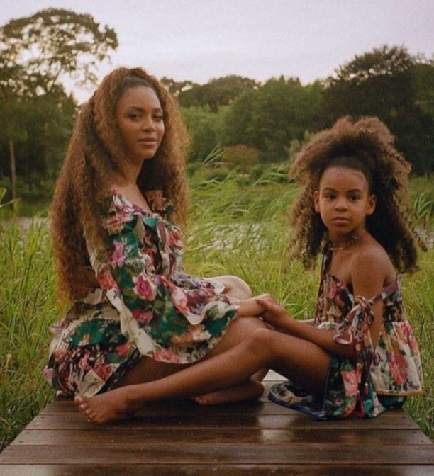 two women sitting on the ground in front of some tall grass and trees, one is wearing a floral dress