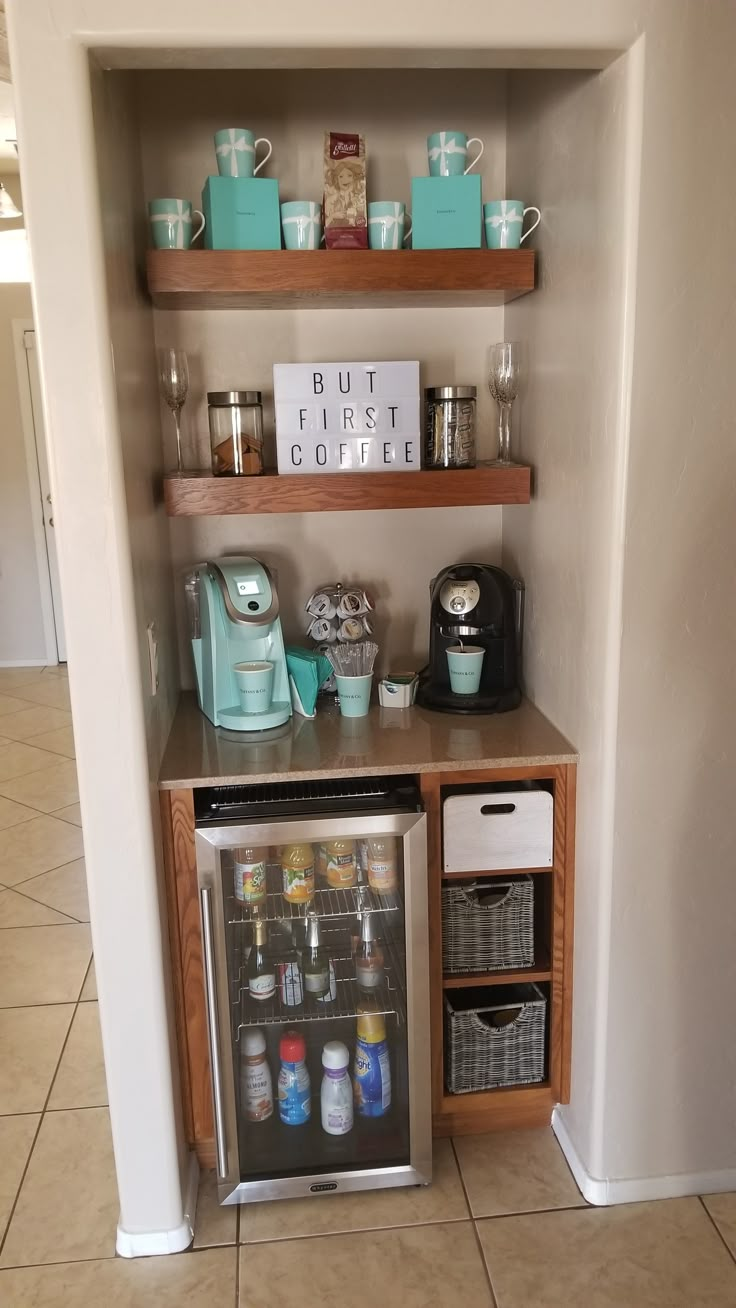 a small refrigerator in the corner of a kitchen next to a shelf with cups and mugs on it