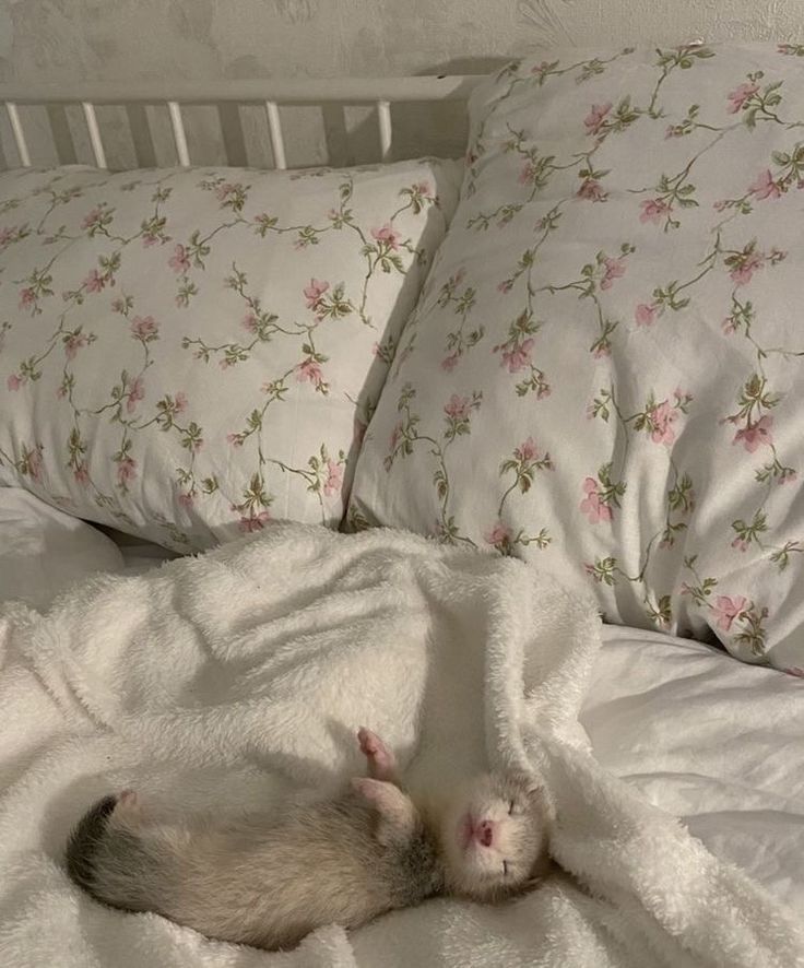 a baby animal sleeping on top of a white bed covered in blankets and pillows with pink flowers