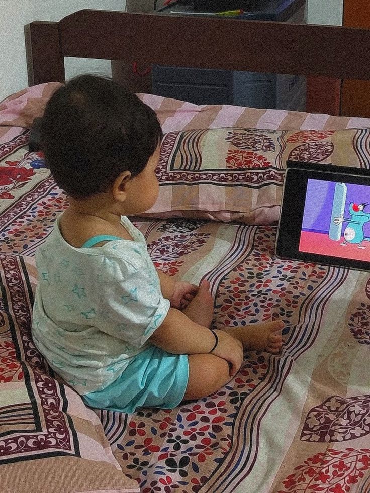 a small child sitting on top of a bed next to a tablet computer monitor with an animated character on it