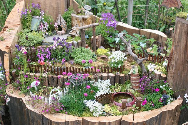 an outdoor garden with lots of flowers and plants on it's sides, in the middle of a wooden fence