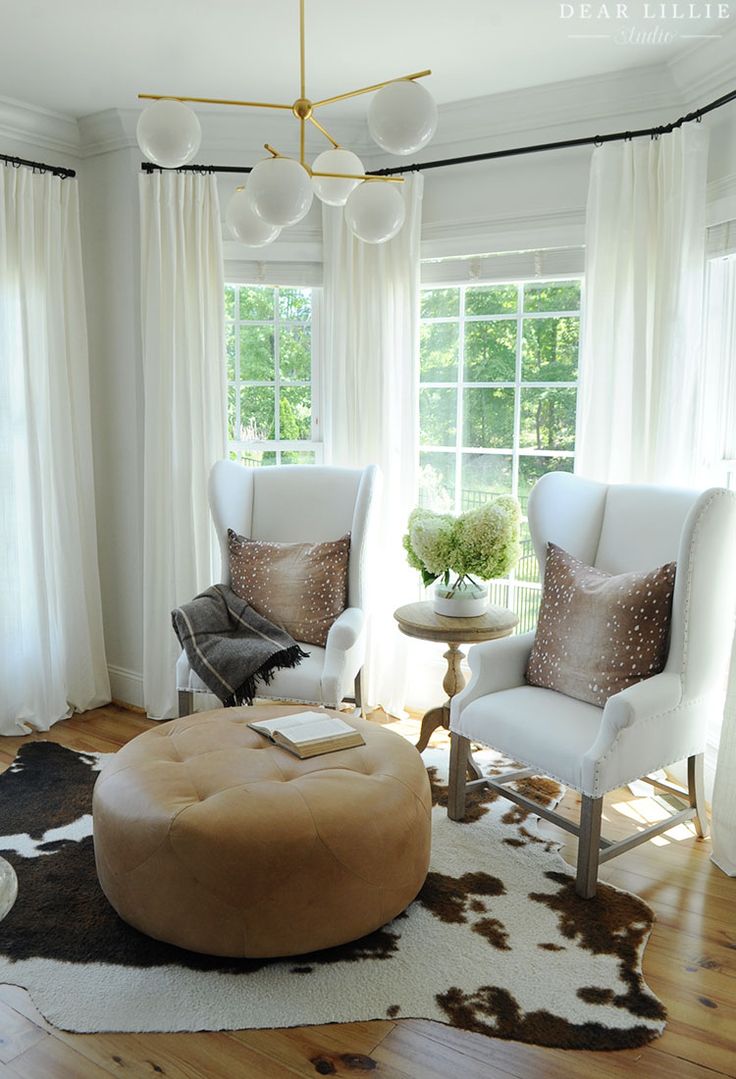 a living room with two white chairs and a round ottoman in front of the window