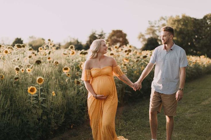 a pregnant couple holding hands and walking through a field of sunflowers