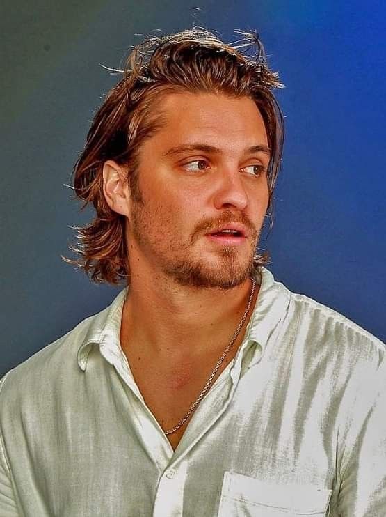 a man with long hair wearing a white shirt and looking off to the side while standing in front of a blue background