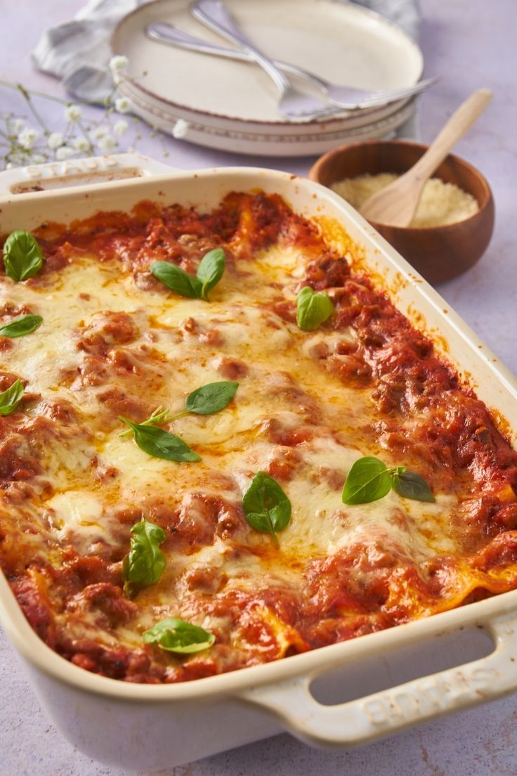 a casserole dish with meat, cheese and basil on the table next to other dishes