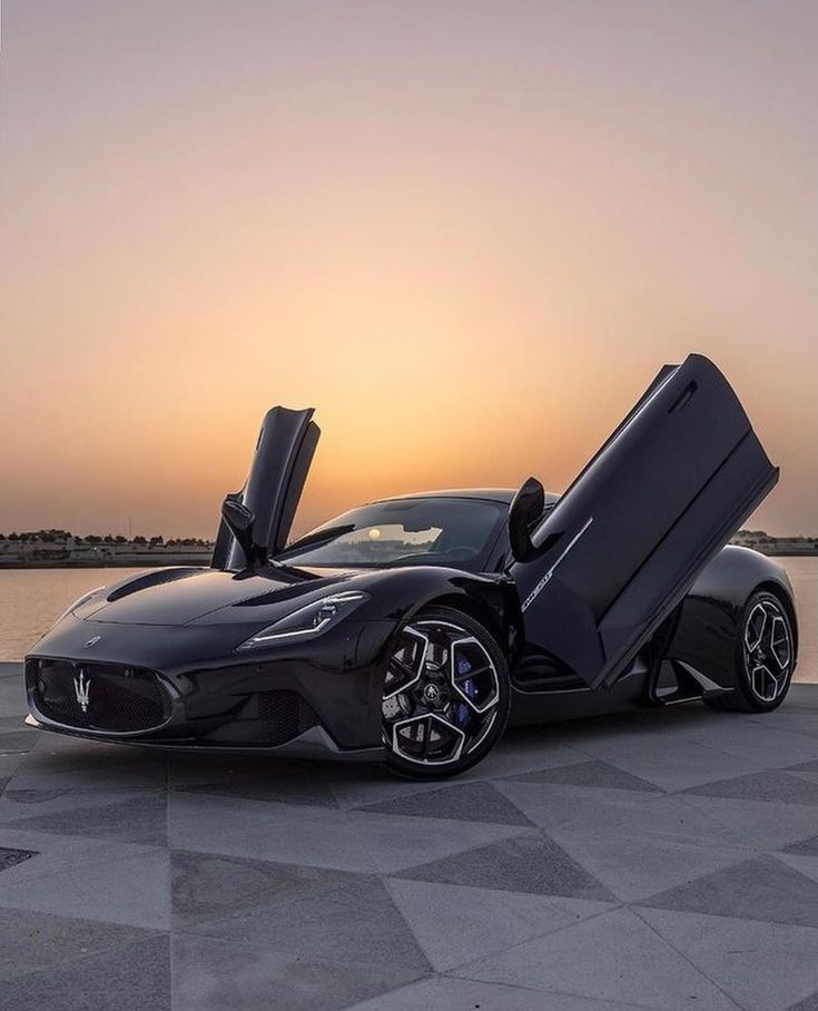 a black sports car with its doors open sitting in front of the water at sunset