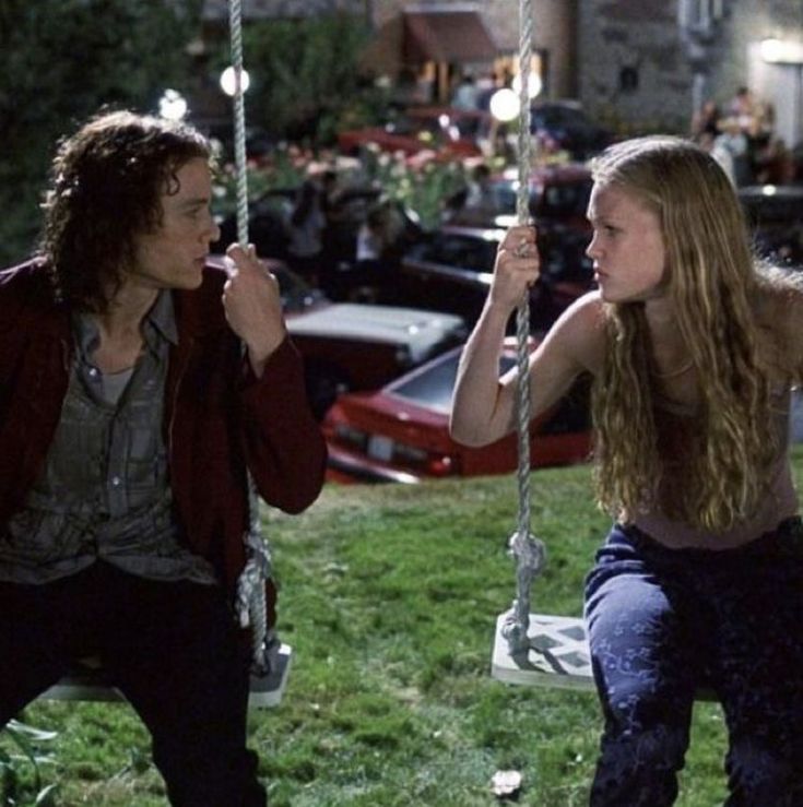 a man and woman sitting on swings in front of parked cars, talking to each other