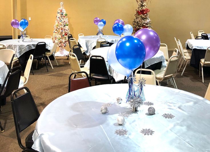 a room filled with tables and chairs covered in white tablecloths topped with blue and purple balloons