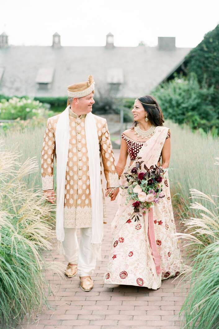 a bride and groom holding hands walking down a path in front of tall grass with flowers