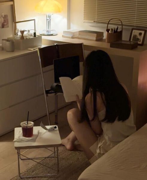 a woman is sitting on the bed reading a book and drinking from a red cup