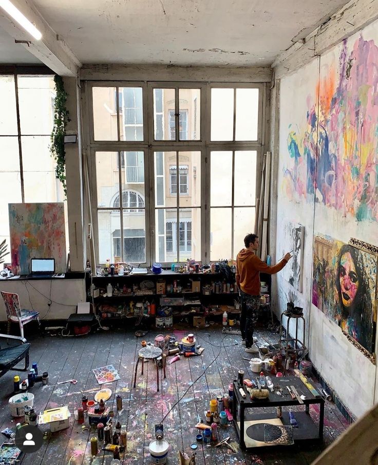 a man standing in an art studio with lots of paintings on the wall and floor