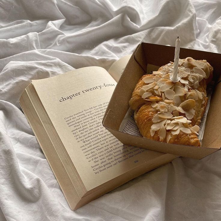 an open book on a bed next to a pastry in a box with icing