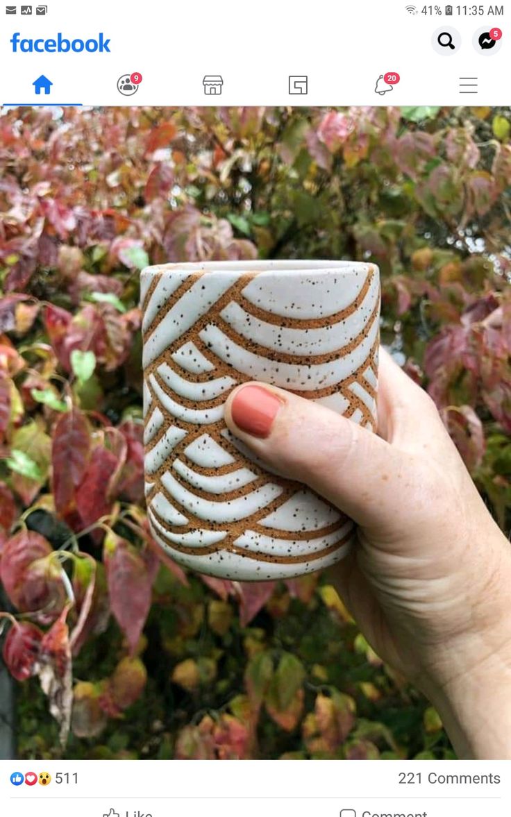 a hand holding a white and brown coffee cup in front of some trees with red leaves