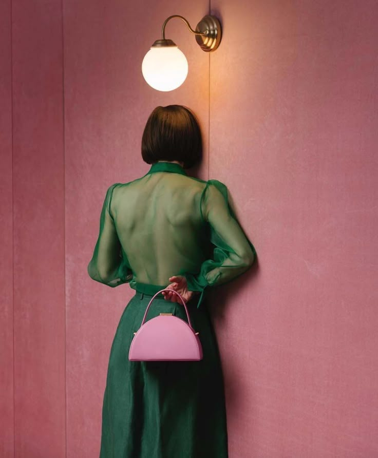 a woman in a green dress is holding a pink purse and standing against a wall