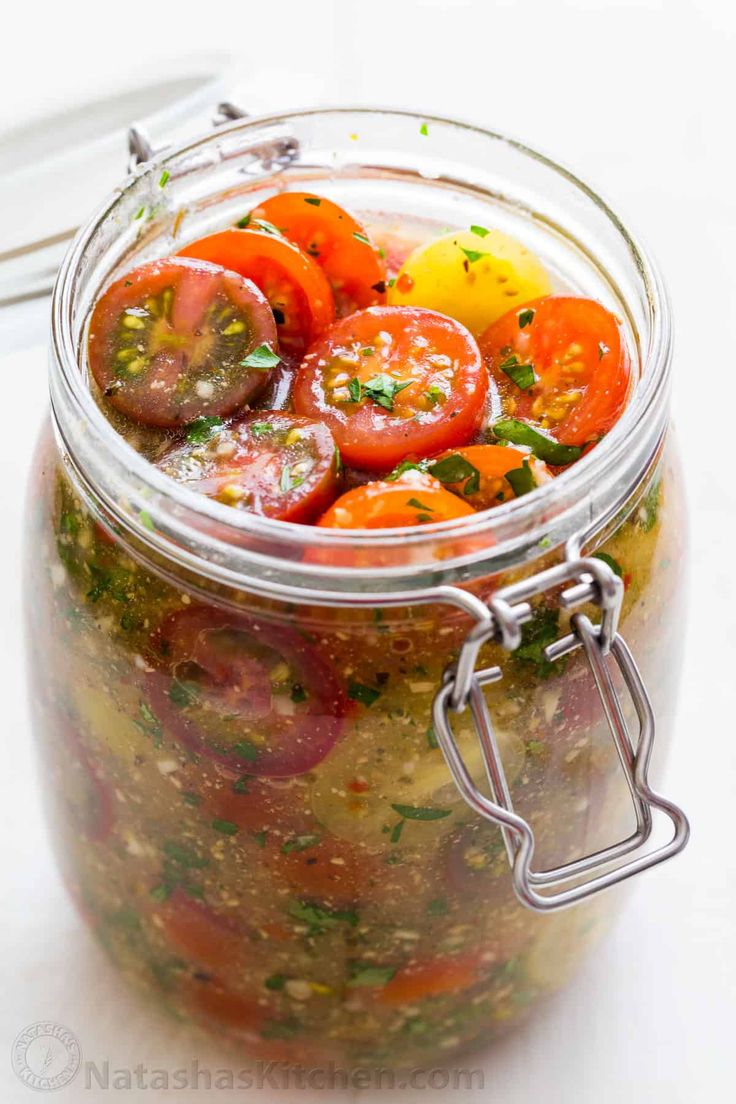 a glass jar filled with lots of different types of tomatoes and herbs on top of a white table