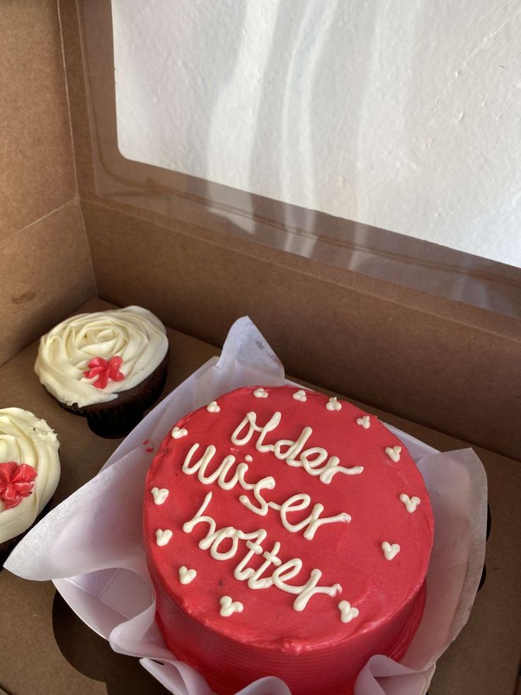 a red cake with white frosting in a box next to two cupcakes