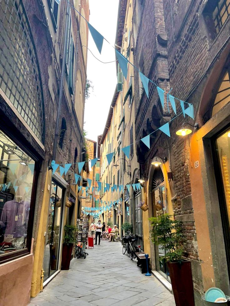 an alley way with people walking down it and blue flags hanging from the buildings above