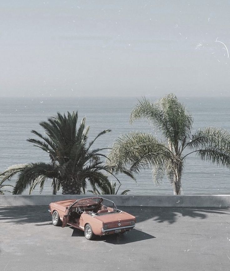 an old pink car parked in front of palm trees and the ocean behind it,