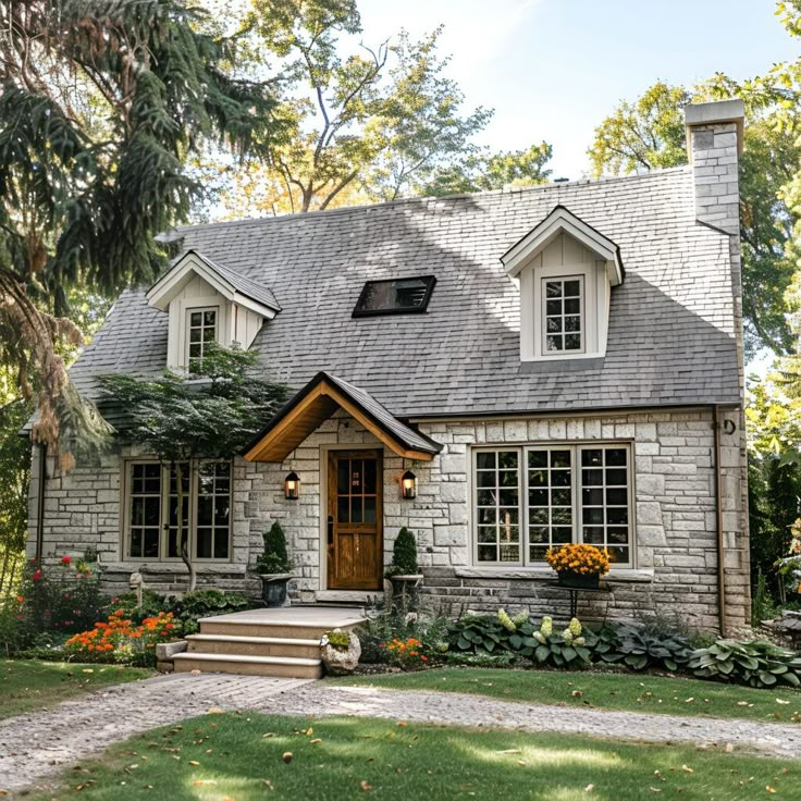 This charming cottage features a classic stone exterior, steep roof with dormer windows, and a welcoming wooden front door. Surrounded by a lush, landscaped garden with blooming flowers and greenery, the large windows brighten the interior. A stone pathway leads to the entrance, enhancing the cottage's quaint and inviting atmosphere, making it the perfect Pinterest-worthy retreat. Country House Small Cottage Style, Single Story Houses Exterior, Tutor Homes Exterior English Cottages, Cute House Entrance, Front Gable House, Small Stone Cottage House Plans, Older Homes Exterior, Cottage With Dormers, Small Cottage Homes Exterior