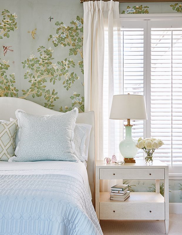 a white bed sitting under a window next to a nightstand with a lamp on top of it