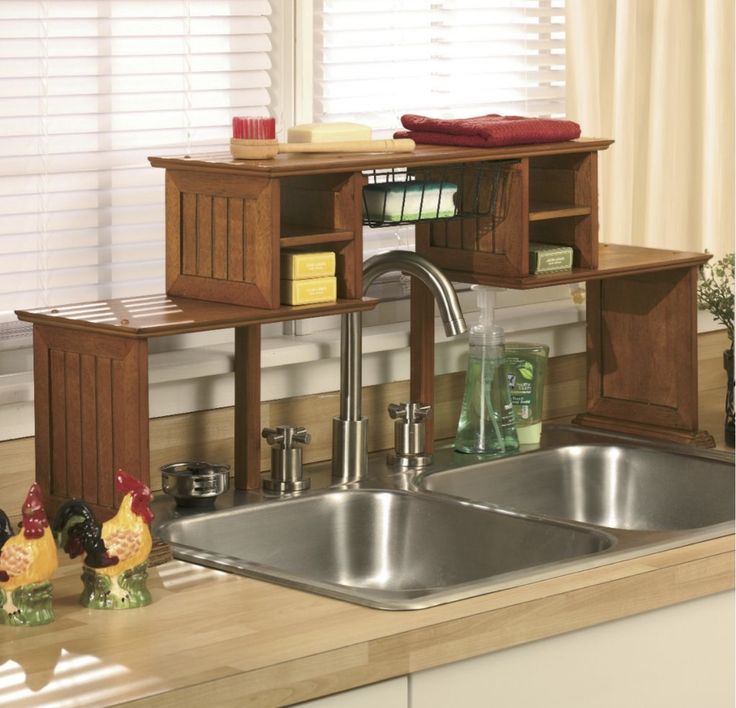 a kitchen sink sitting under a window next to a wooden shelf filled with clutter