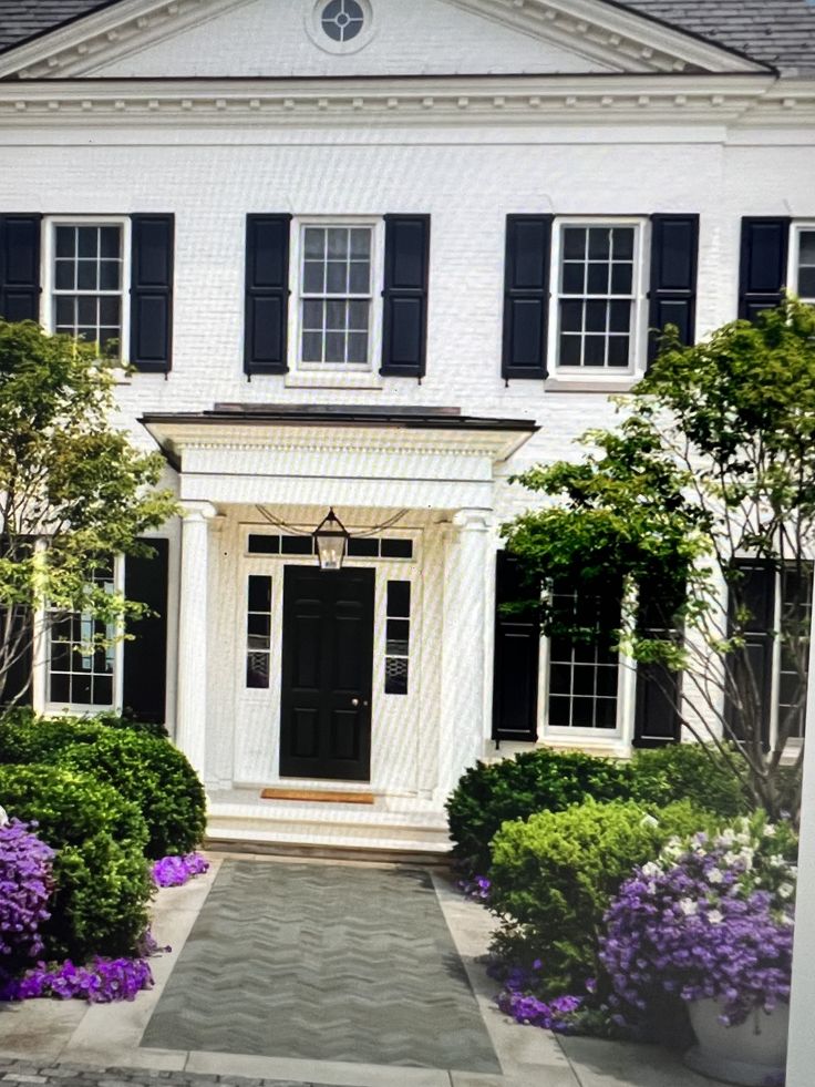 a large white house with black shutters and flowers in the front yard on a sunny day