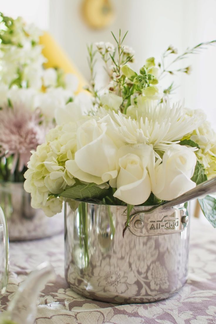 white flowers are in a silver vase on a table with other floral arrangements behind it