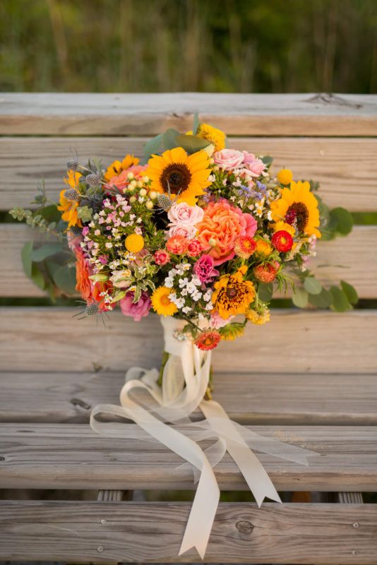 a bouquet of sunflowers and other flowers on a wooden bench with white ribbon