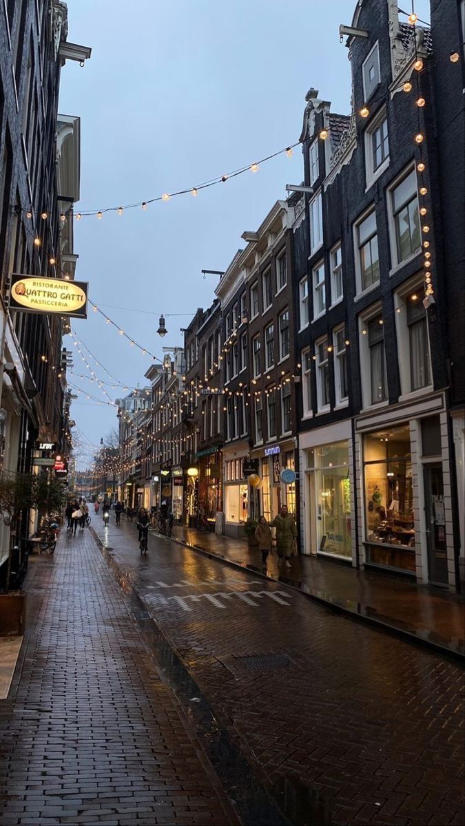 an empty city street with buildings and lights strung over the sidewalks on a rainy day