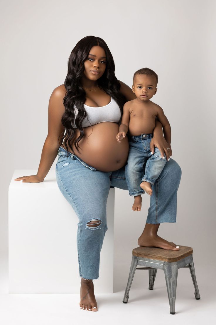 a pregnant woman sitting on top of a stool next to a baby boy wearing ripped jeans