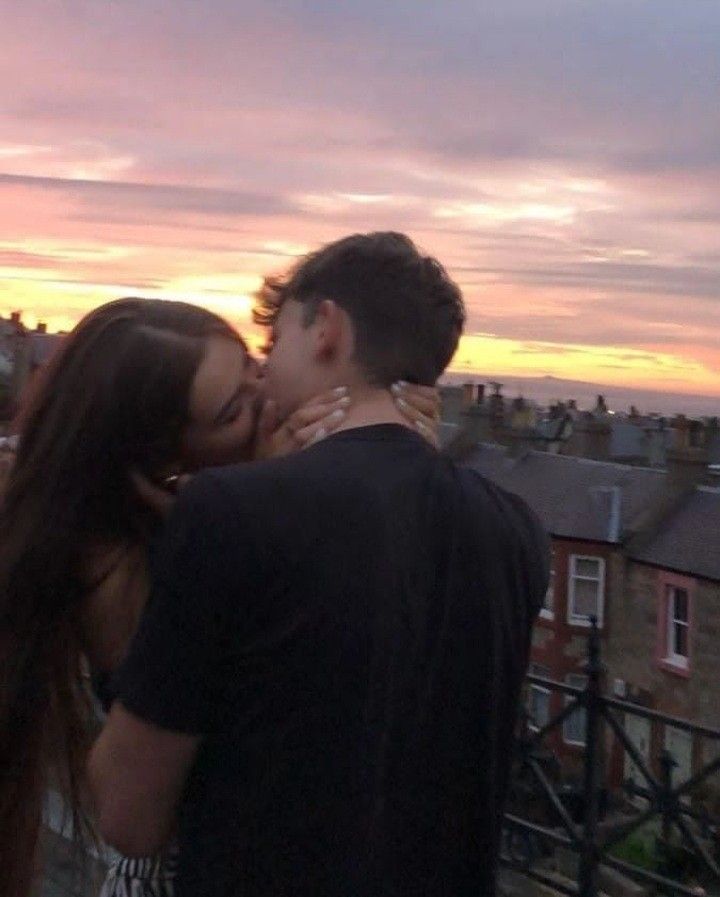 a man and woman kissing in front of a sunset over rooftops with buildings behind them