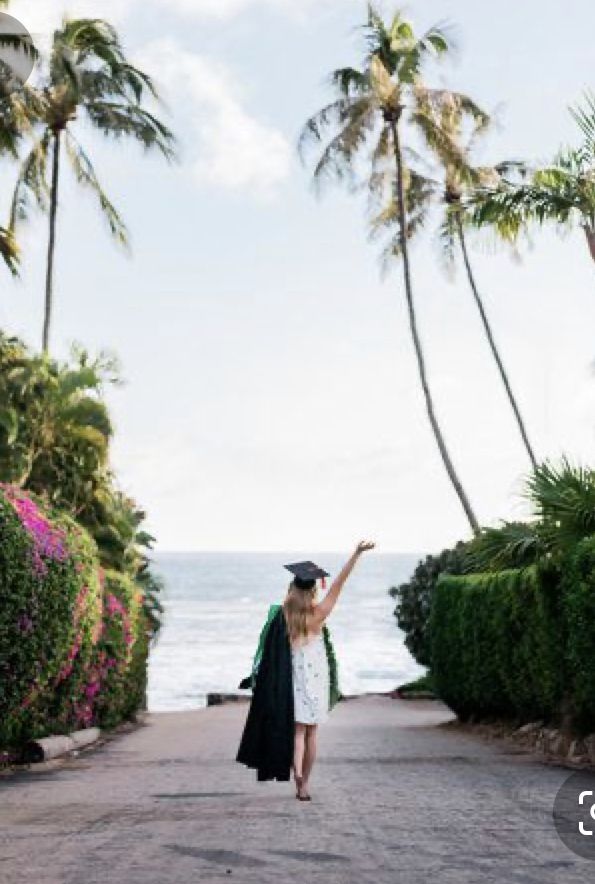 a woman in graduation gown and cap walking down a path towards the ocean with palm trees