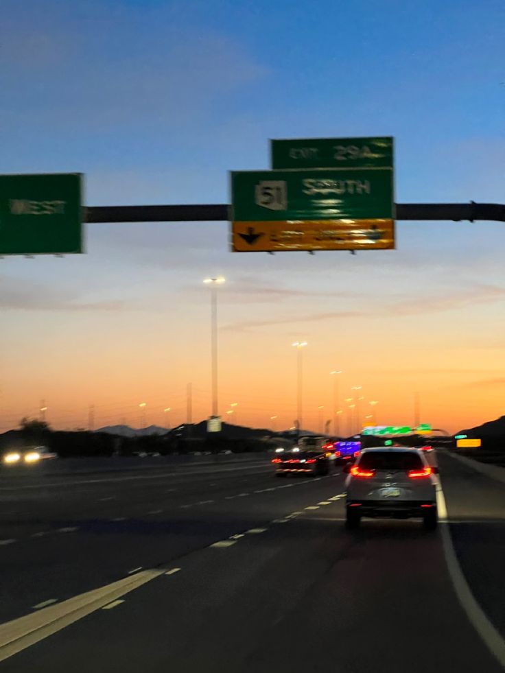 two green and white street signs on a pole above a highway at dusk with traffic lights