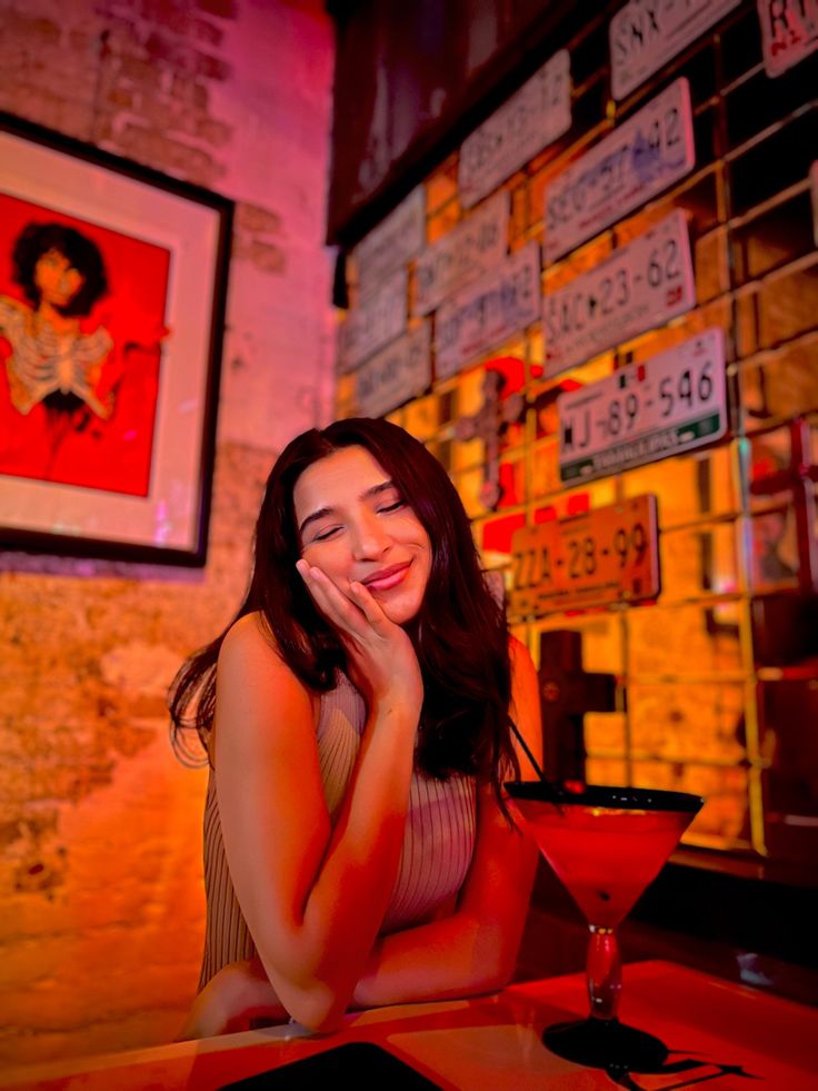 a woman sitting at a table with a drink in front of her and signs on the wall behind her