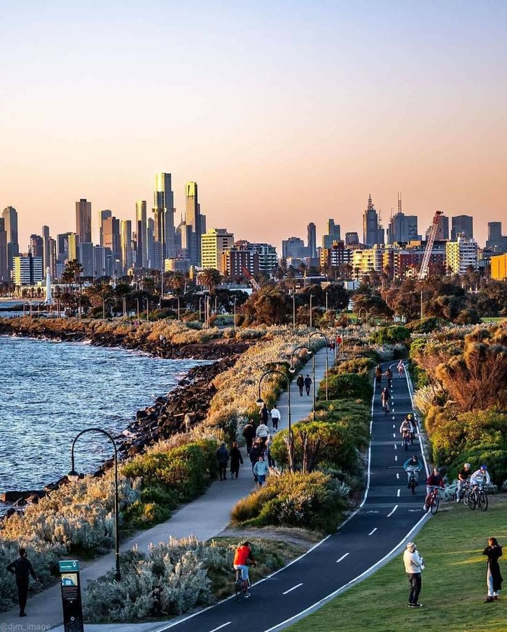 people walking and biking along a path near the water with city skyline in the background