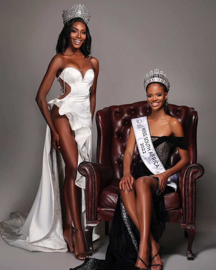 two beautiful women sitting next to each other in front of a brown chair and one wearing a tiara