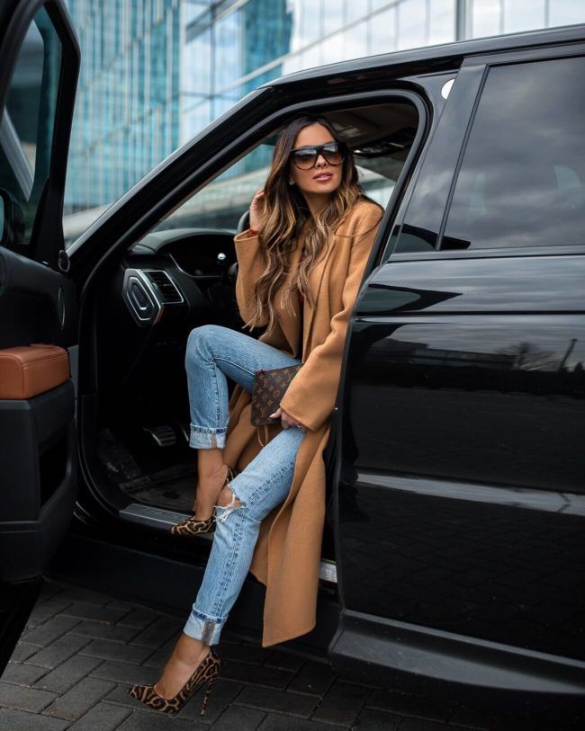 a woman sitting in the door of a black car wearing leopard print shoes and a camel coat