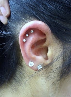 a woman with black hair has three piercings on her ear and behind her ear is a white diamond