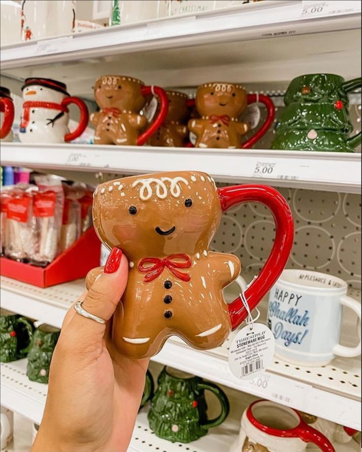 a hand holding a gingerbread man ornament in front of shelves with mugs