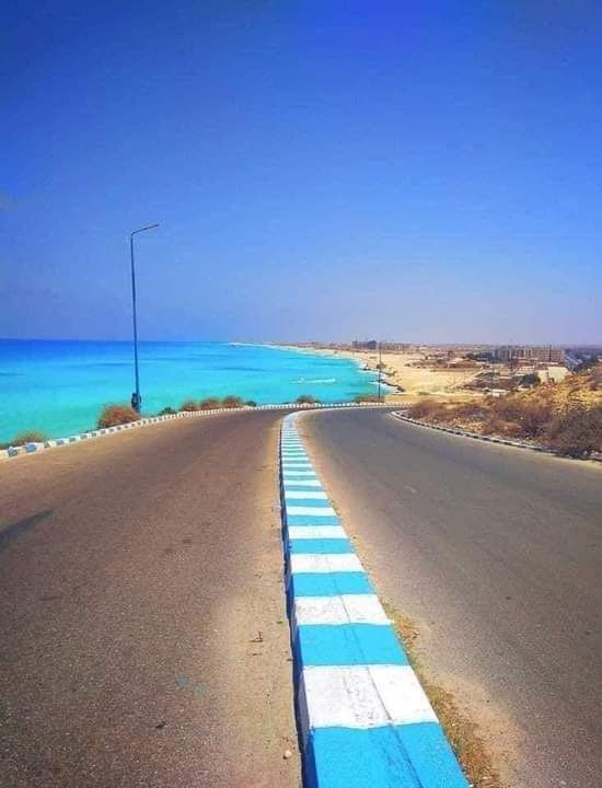 an empty road next to the ocean with blue water in the backgrouds