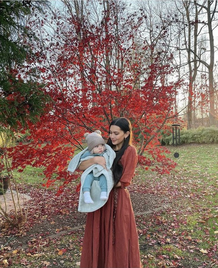 a woman holding a baby in her arms near a tree with red leaves on it