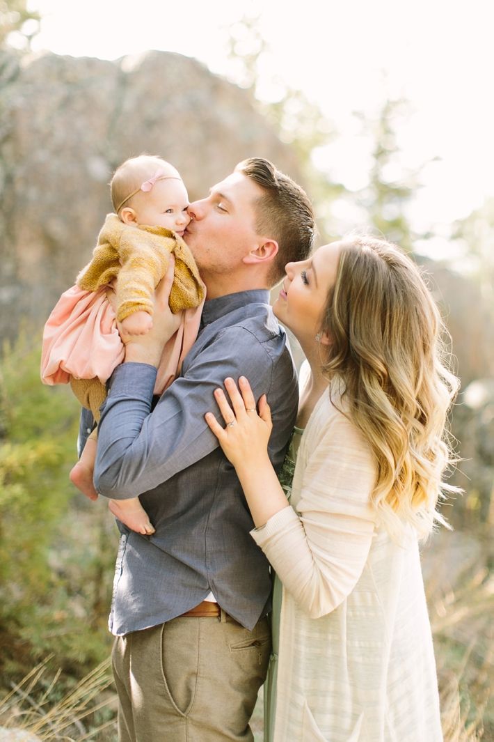 a man and woman kissing while holding a baby in the air with trees behind them