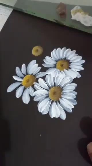 someone is drawing daisies on a piece of black paper with white and yellow flowers