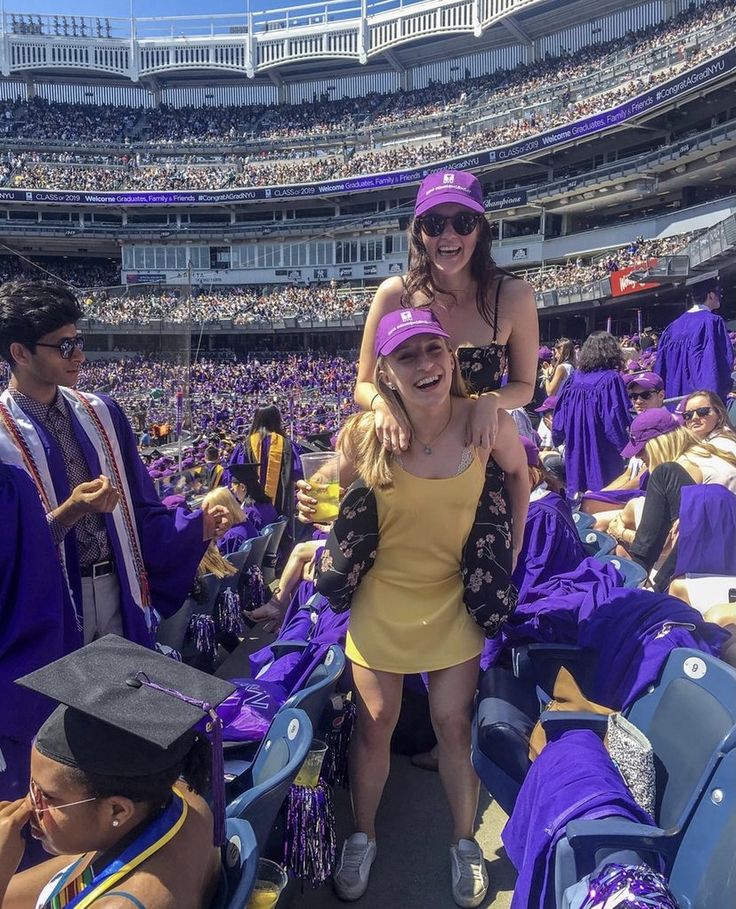 two women in graduation gowns sitting on the stands at a stadium with other people