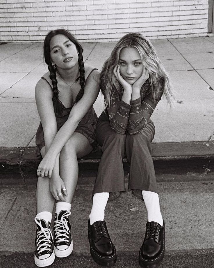 two young women sitting on the curb with their feet up against each other's legs